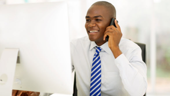 image of man on phone at desk looking at computer screen smiling